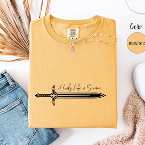 Puede incluir: Camiseta color mostaza con un gr&aacute;fico de espada negra y el texto "it looks like a Simon". La camiseta se combina con vaqueros azules, zapatos blancos y un collar. El color es mostaza.