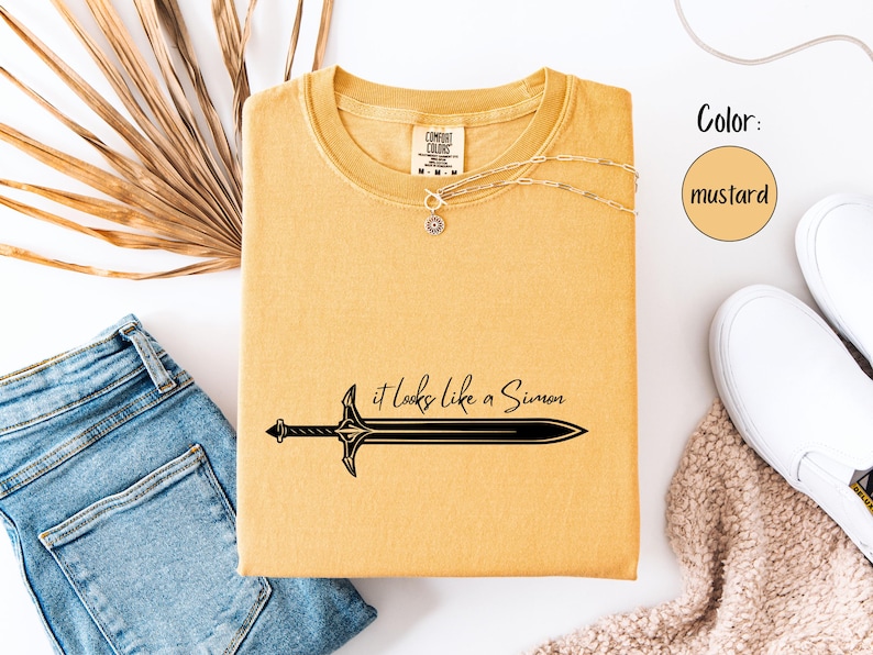 Puede incluir: Camiseta color mostaza con un gr&aacute;fico de espada negra y el texto "it looks like a Simon". La camiseta se combina con vaqueros azules, zapatos blancos y un collar. El color es mostaza.
