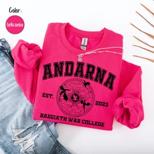 May include: A hot pink sweatshirt with the word "ANDARNA" in black block letters. Below the word is a circular graphic with the text "BASGIATH WAR COLLEGE" and the year "2023". A silver necklace is draped across the sweatshirt.