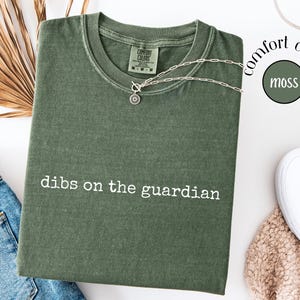 May include: A moss green short-sleeve t-shirt with a crew neckline and the white text "dibs on the guardian" printed on the chest. The shirt features a Comfort Colors brand label at the inner neck and a vintage-style washed fabric finish.