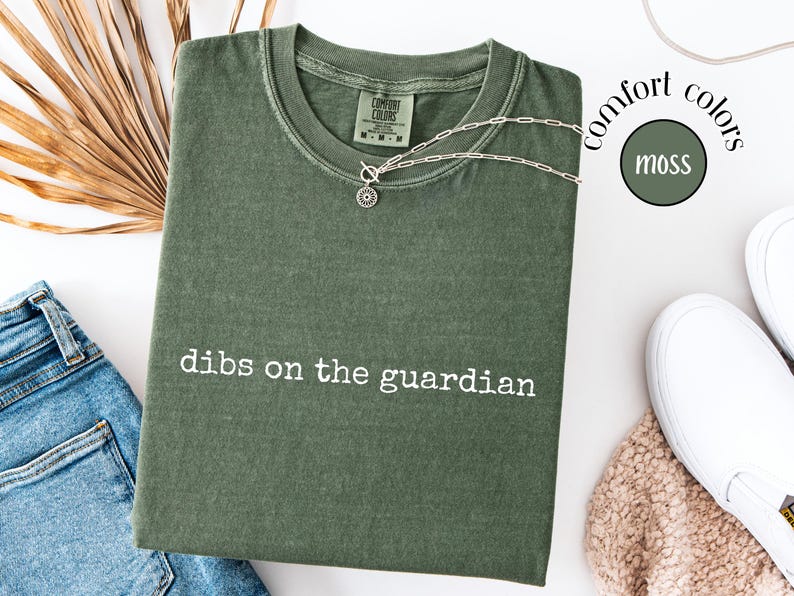 May include: A moss green short-sleeve t-shirt with a crew neckline and the white text "dibs on the guardian" printed on the chest. The shirt features a Comfort Colors brand label at the inner neck and a vintage-style washed fabric finish.