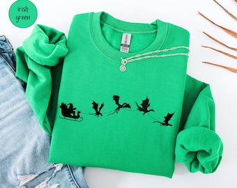 May include: Irish green sweatshirt with a black graphic of Santa Claus in a sleigh pulled by dragons. The sweatshirt has a crew neck and long sleeves. A silver necklace with a pendant is draped on the sweatshirt. The color is labeled as "Irish green".