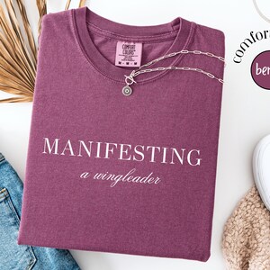 Op de afbeelding: Een bessenkleurig T-shirt met de woorden "MANIFESTING a wingleader" in het wit gedrukt. Een zilveren ketting met een ronde hanger ligt op het shirt. Het shirt is opgevouwen naast een spijkerbroek en witte sneakers.
