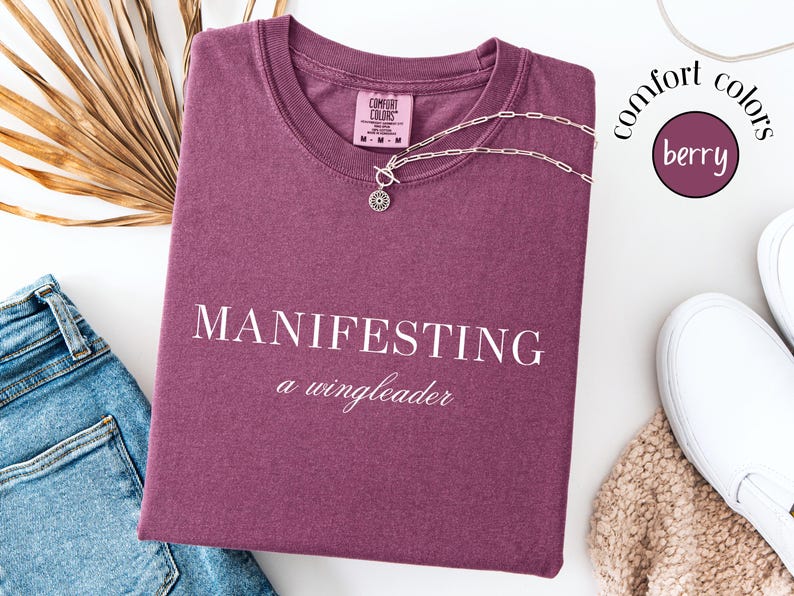 Op de afbeelding: Een bessenkleurig T-shirt met de woorden "MANIFESTING a wingleader" in het wit gedrukt. Een zilveren ketting met een ronde hanger ligt op het shirt. Het shirt is opgevouwen naast een spijkerbroek en witte sneakers.