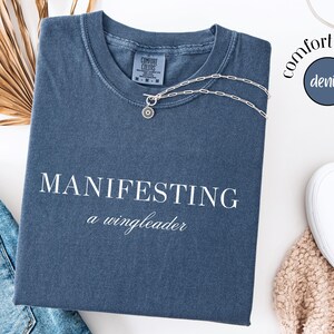 Op de afbeelding: Een denim blauw t-shirt met de witte tekst "MANIFESTING a wingleader". Een zilveren ketting met een ronde hanger ligt op het shirt. Het shirt is opgevouwen naast een spijkerbroek en witte instappers.