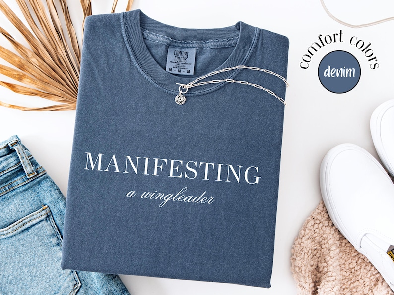 Op de afbeelding: Een denim blauw t-shirt met de witte tekst "MANIFESTING a wingleader". Een zilveren ketting met een ronde hanger ligt op het shirt. Het shirt is opgevouwen naast een spijkerbroek en witte instappers.