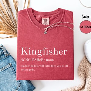 May include: Crimson-colored t-shirt with the word "Kingfisher" printed in white, along with a definition. A silver necklace with a pendant is on the shirt. The shirt is paired with blue jeans and white sneakers.