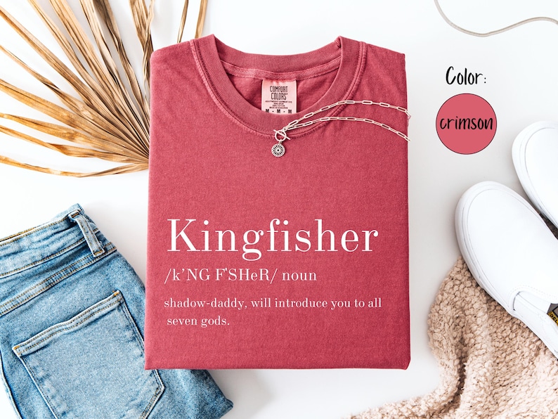 May include: Crimson-colored t-shirt with the word "Kingfisher" printed in white, along with a definition. A silver necklace with a pendant is on the shirt. The shirt is paired with blue jeans and white sneakers.