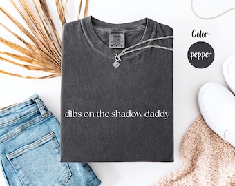 May include: Pepper-colored t-shirt with the phrase "dibs on the shadow daddy" in white. A silver necklace with a pendant rests on the shirt. The image includes blue jeans, white shoes, and a dried plant.