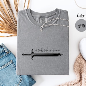 Puede incluir: Camiseta gris con un gr&aacute;fico de espada negra y la frase "it looks like a Simon". Un collar plateado con un colgante circular est&aacute; sobre la camiseta. La camiseta est&aacute; junto a unos vaqueros azules y zapatillas blancas.