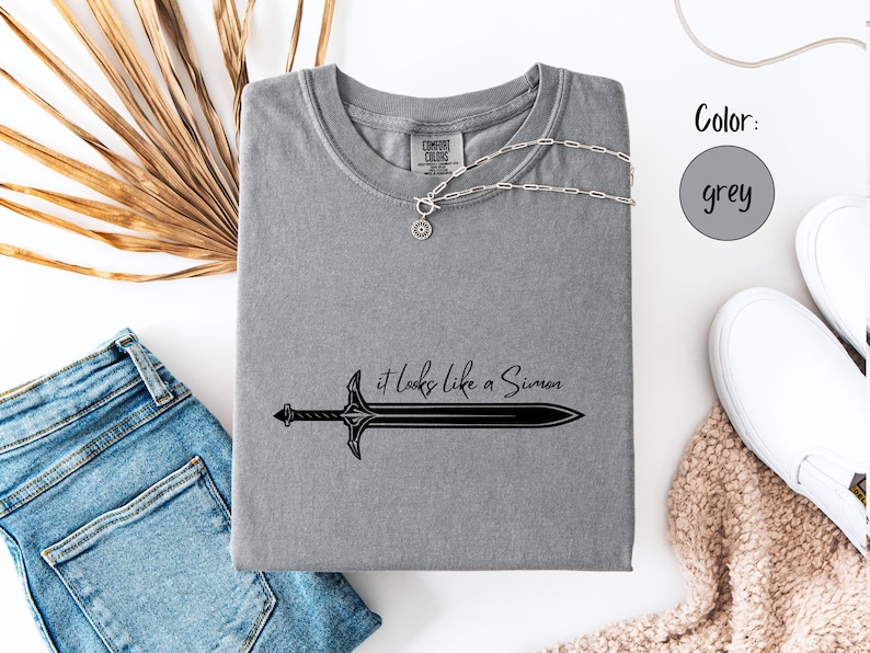 Puede incluir: Camiseta gris con un gr&aacute;fico de espada negra y la frase "it looks like a Simon". Un collar plateado con un colgante circular est&aacute; sobre la camiseta. La camiseta est&aacute; junto a unos vaqueros azules y zapatillas blancas.