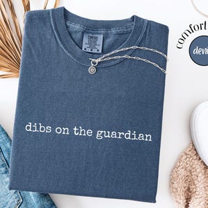 May include: A denim blue short-sleeved t-shirt with the text "dibs on the guardian" embroidered in white across the chest. The shirt features a crew neckline and a Comfort Colors brand label at the inner collar, paired with a silver chain necklace.