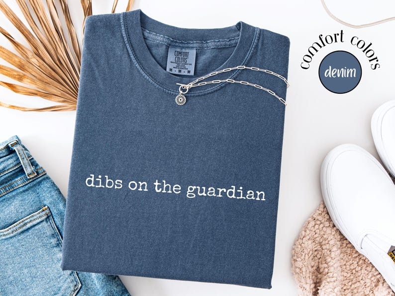 May include: A denim blue short-sleeved t-shirt with the text "dibs on the guardian" embroidered in white across the chest. The shirt features a crew neckline and a Comfort Colors brand label at the inner collar, paired with a silver chain necklace.
