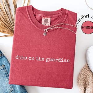 May include: A crimson short-sleeve t-shirt with the white text "dibs on the guardian" printed on the front. The shirt features a crew neckline and a Comfort Colors brand label at the inner collar. A silver-toned chain necklace rests on the shirt.
