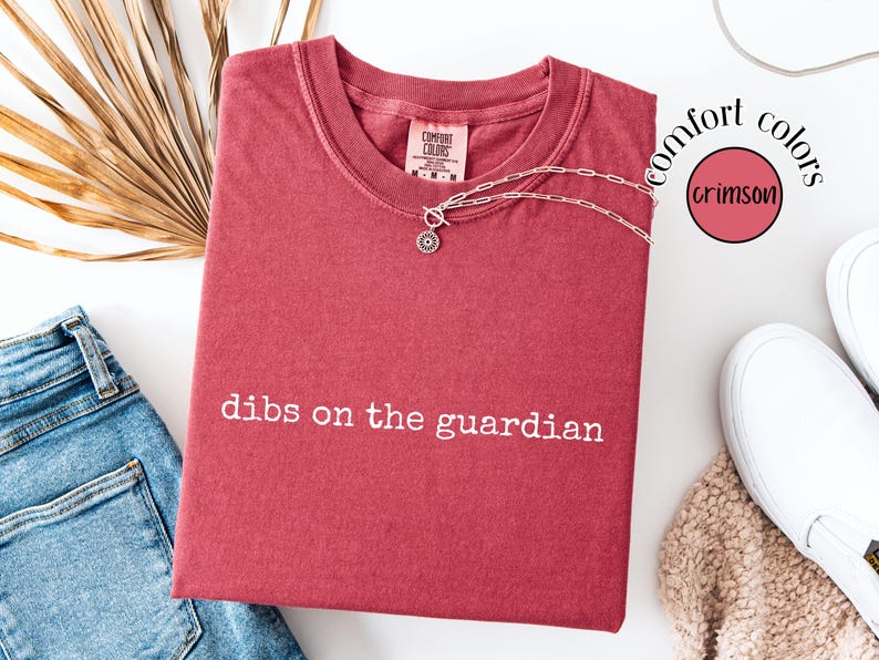 May include: A crimson short-sleeve t-shirt with the white text "dibs on the guardian" printed on the front. The shirt features a crew neckline and a Comfort Colors brand label at the inner collar. A silver-toned chain necklace rests on the shirt.