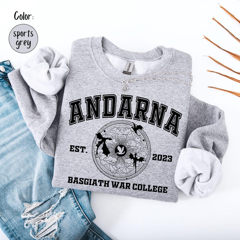 May include: A sports grey sweatshirt with the word "ANDARNA" in black block letters. Below the word is a circular graphic with the text "EST. 2023" and "BASGIATH WAR COLLEGE". The sweatshirt is folded on top of blue jeans.