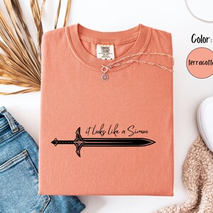 Puede incluir: Camiseta color terracota con un gr&aacute;fico de espada negra y la frase "it looks like a Simon". La camiseta se combina con vaqueros azules, zapatillas blancas y un collar plateado. El color se indica en un c&iacute;rculo.