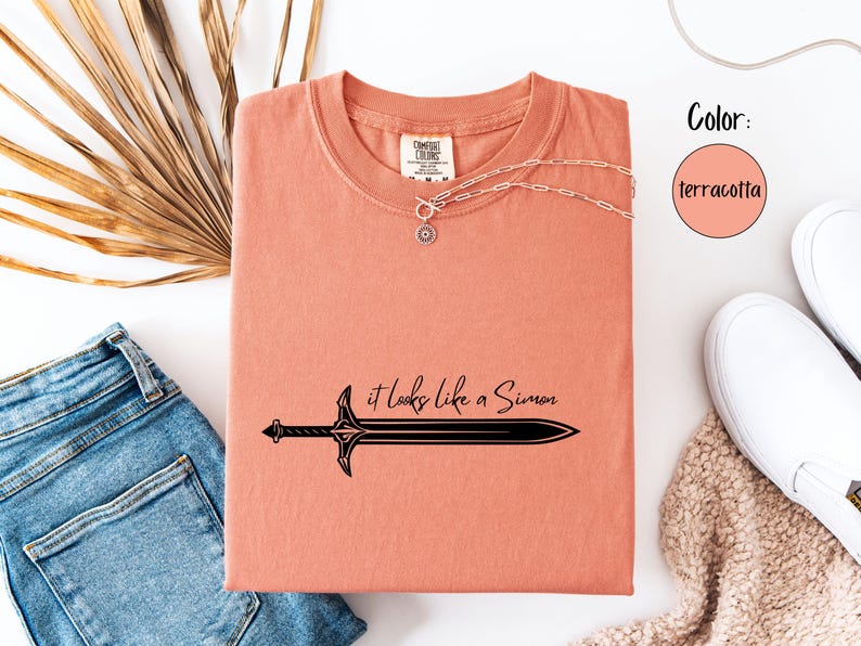 Puede incluir: Camiseta color terracota con un gr&aacute;fico de espada negra y la frase "it looks like a Simon". La camiseta se combina con vaqueros azules, zapatillas blancas y un collar plateado. El color se indica en un c&iacute;rculo.