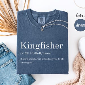 May include: A denim-colored t-shirt with the word "Kingfisher" printed in white, along with a definition. A silver necklace with a pendant rests on the shirt. The shirt is paired with blue jeans and white sneakers.