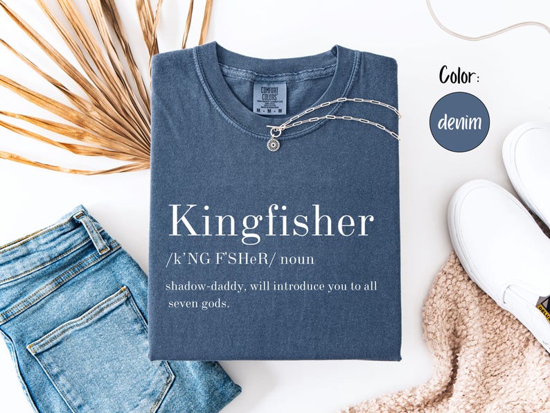 May include: A denim-colored t-shirt with the word "Kingfisher" printed in white, along with a definition. A silver necklace with a pendant rests on the shirt. The shirt is paired with blue jeans and white sneakers.