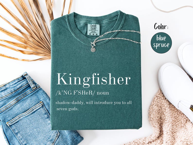 May include: A blue spruce t-shirt with the word "Kingfisher" printed in white, along with a definition. A silver necklace and blue jeans are next to the shirt. The shirt is folded on a white surface.