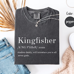 Könnte beinhalten: Ein dunkelgraues T-Shirt mit dem Wort "Kingfisher" in Weiß aufgedruckt, zusammen mit einer Definition. Eine silberne Halskette liegt auf dem Shirt. Das Shirt wird mit blauen Jeans, weißen Schuhen und einem beigen Schal präsentiert.