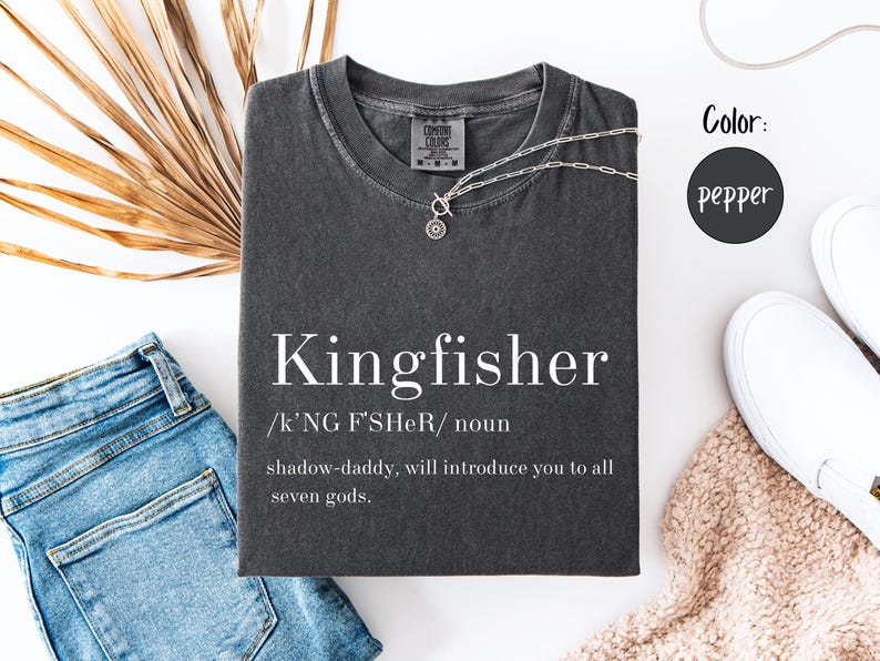 May include: A pepper-colored t-shirt with the word "Kingfisher" printed in white, along with a definition. A silver necklace rests on the shirt. The shirt is displayed with blue jeans, white shoes, and a beige scarf.