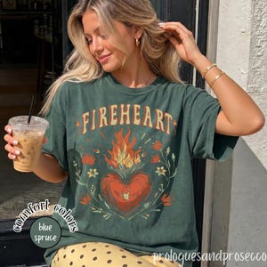 May include: A dark green short-sleeve t-shirt featuring the text FIREHEART in a curved font above a graphic of a flaming red heart surrounded by orange and red flowers. The shirt has a crew neckline and a relaxed fit with a vintage-style wash.