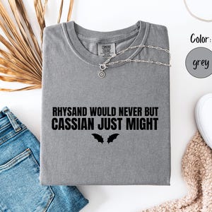 Könnte beinhalten: Ein graues T-Shirt mit dem Text "RHYSAND WOULD NEVER BUT CASSIAN JUST MIGHT" und zwei Fledermaus-Silhouetten. Eine silberne Halskette liegt über dem Shirt. Das Shirt liegt auf einer weißen Oberfläche mit blauen Jeans und weißen Turnschuhen.