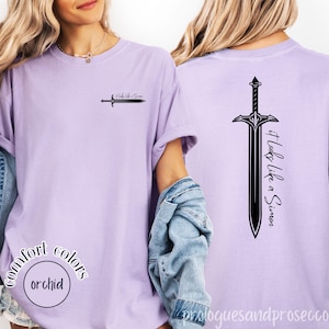 Può includere: T-shirt lilla chiaro con la scritta "It Looks Like a Simon" e un disegno di spada sul davanti e sul retro. La maglietta presenta anche le parole "Comfort Colors" e "orchid".