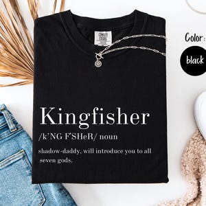 May include: Black t-shirt with the word "Kingfisher" in white, along with a definition. A silver necklace is draped across the shirt. The color black is indicated in a circle. The shirt is folded on a white surface.