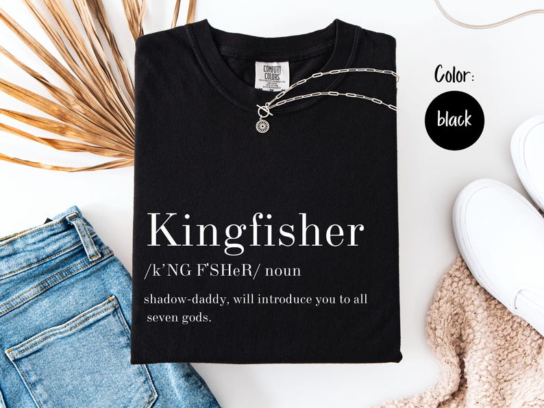 May include: Black t-shirt with the word "Kingfisher" in white, along with a definition. A silver necklace is draped across the shirt. The color black is indicated in a circle. The shirt is folded on a white surface.