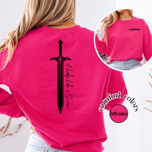 May include: A bright pink sweatshirt with a black sword graphic and the text "It looks like a Simon" on the back. The front of the sweatshirt has a smaller sword graphic. The words "comfort colors" and "heliconia" are also visible.