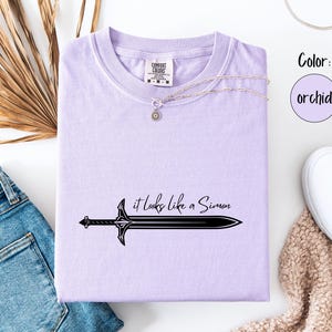 Puede incluir: Camiseta color orqu&iacute;dea con un gr&aacute;fico de espada negra y el texto "it looks like a Simon". La camiseta se combina con vaqueros azules, zapatillas blancas y un collar plateado. El color se indica como orqu&iacute;dea.