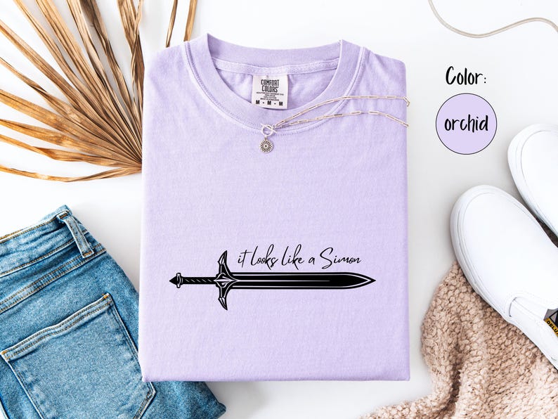 Puede incluir: Camiseta color orqu&iacute;dea con un gr&aacute;fico de espada negra y el texto "it looks like a Simon". La camiseta se combina con vaqueros azules, zapatillas blancas y un collar plateado. El color se indica como orqu&iacute;dea.