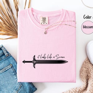 Puede incluir: Camiseta rosa claro con el texto "it looks like a Simon" y un dise&ntilde;o de espada negra. La camiseta se muestra con vaqueros azules, un collar y zapatos blancos. El color es blossom.