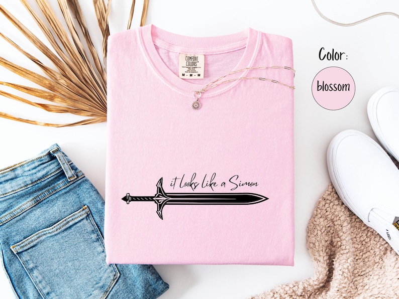 Puede incluir: Camiseta rosa claro con el texto "it looks like a Simon" y un dise&ntilde;o de espada negra. La camiseta se muestra con vaqueros azules, un collar y zapatos blancos. El color es blossom.