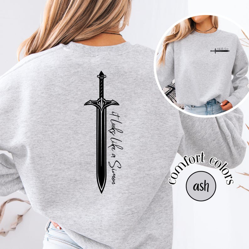 May include: Light grey sweatshirt with a black sword graphic on the back and a smaller sword graphic on the front. The text "It looks like a Simon" is printed vertically on the back. The sweatshirt is labeled "comfort colors" and "ash".