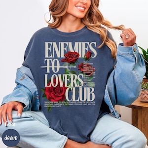 以下が含まれることがあります： 「ENEMIES TO LOVERS CLUB」の文字と赤いバラのイラストが描かれたデニムブルーのスウェットシャツ。テキストにはテーマの説明が含まれています。女性がスウェットシャツを着ています。
