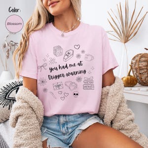 Puede incluir: Camiseta rosa claro con el texto negro "you had me at trigger warning". La camiseta presenta varias ilustraciones de línea negra, incluyendo un ataúd, libros, corazones y una calavera. Color: Blossom.
