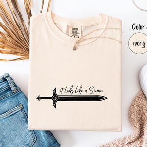 Puede incluir: Camiseta de color marfil con el texto "it looks like a Simon" y un gr&aacute;fico de espada negra. La camiseta se muestra con vaqueros azules y zapatillas blancas sobre una superficie blanca.