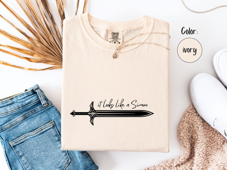 Puede incluir: Camiseta de color marfil con el texto "it looks like a Simon" y un gr&aacute;fico de espada negra. La camiseta se muestra con vaqueros azules y zapatillas blancas sobre una superficie blanca.