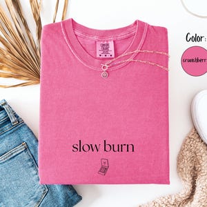 May include: A pink t-shirt with the words "slow burn" printed on the front, along with a small graphic. A silver necklace is draped across the shirt. The shirt is paired with blue jeans and white sneakers.