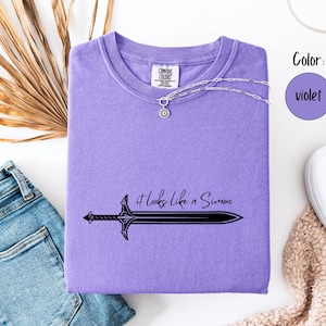 Puede incluir: Una camiseta violeta Comfort Colors con el texto "it looks like a Simon" y un gr&aacute;fico de espada. Un collar plateado con un colgante est&aacute; sobre la camiseta. La camiseta est&aacute; doblada sobre una superficie blanca con vaqueros azules y zapatos blancos.
