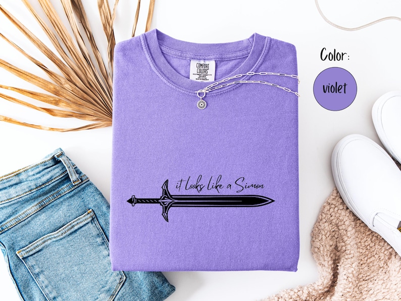 Puede incluir: Una camiseta violeta Comfort Colors con el texto "it looks like a Simon" y un gr&aacute;fico de espada. Un collar plateado con un colgante est&aacute; sobre la camiseta. La camiseta est&aacute; doblada sobre una superficie blanca con vaqueros azules y zapatos blancos.