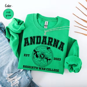 May include: Irish green sweatshirt with the word "ANDARNA" in black block letters. Below the word is a circular graphic with the text "EST. 2023 BASGIATH WAR COLLEGE". A silver necklace rests on the sweatshirt. The sweatshirt is folded next to a pair of blue jeans.