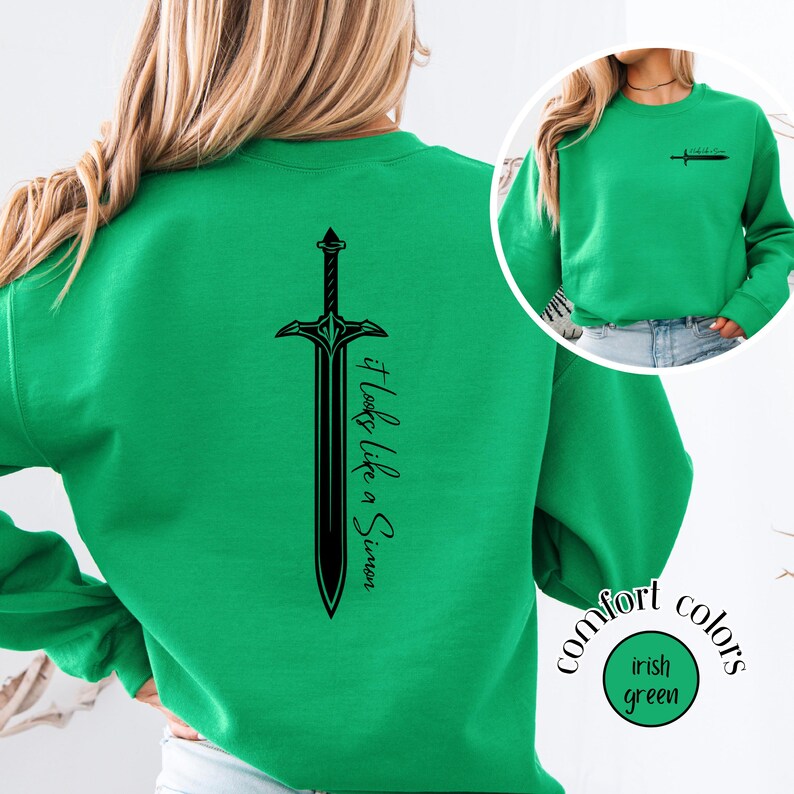 May include: Irish green sweatshirt with a black sword graphic on the back and small sword graphic on the front. The text "It Looks Like a Simon" is printed vertically on the back. The sweatshirt is labeled "Comfort Colors".