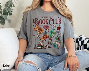 May include: A grey t-shirt featuring the words "North Pole Book Club" and festive illustrations of books, a candle, and a mug. The design has a holiday theme with a cosy, bookish aesthetic. The shirt is a relaxed fit.