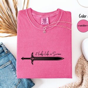 Puede incluir: Una camiseta rosa con el texto "it looks like a Simon" y un gr&aacute;fico de espada negra. Un collar plateado est&aacute; sobre la camiseta. La camiseta est&aacute; junto a unos vaqueros azules y zapatillas blancas.