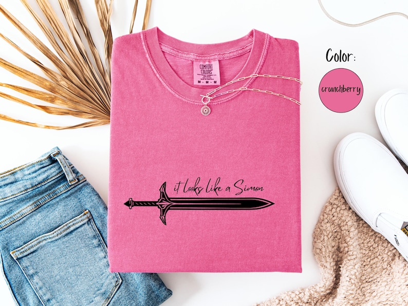 Puede incluir: Una camiseta rosa con el texto "it looks like a Simon" y un gr&aacute;fico de espada negra. Un collar plateado est&aacute; sobre la camiseta. La camiseta est&aacute; junto a unos vaqueros azules y zapatillas blancas.